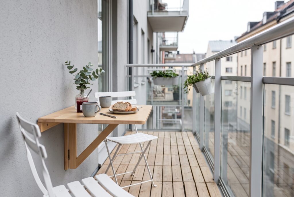 Balcón estrecho con una mesa abatible de madera clara y dos sillas plegables blancas, con el desayuno servido por la mañana