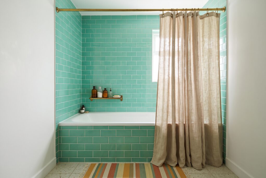 Baño pequeño con cortina de ducha de lino crudo colgada desde una barra a ras de techo y azulejo verde agua en la pared