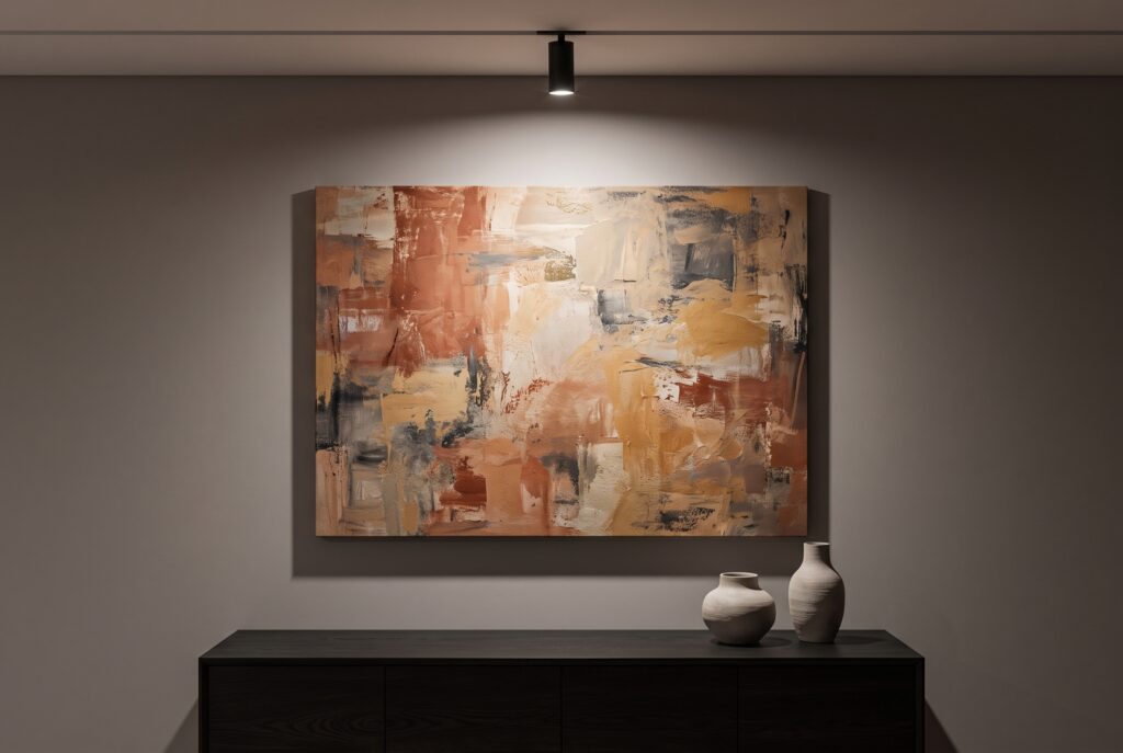 Foco orientable de techo negro iluminando un cuadro abstracto grande en tonos terracota sobre un aparador bajo, con el resto de la sala en penumbra