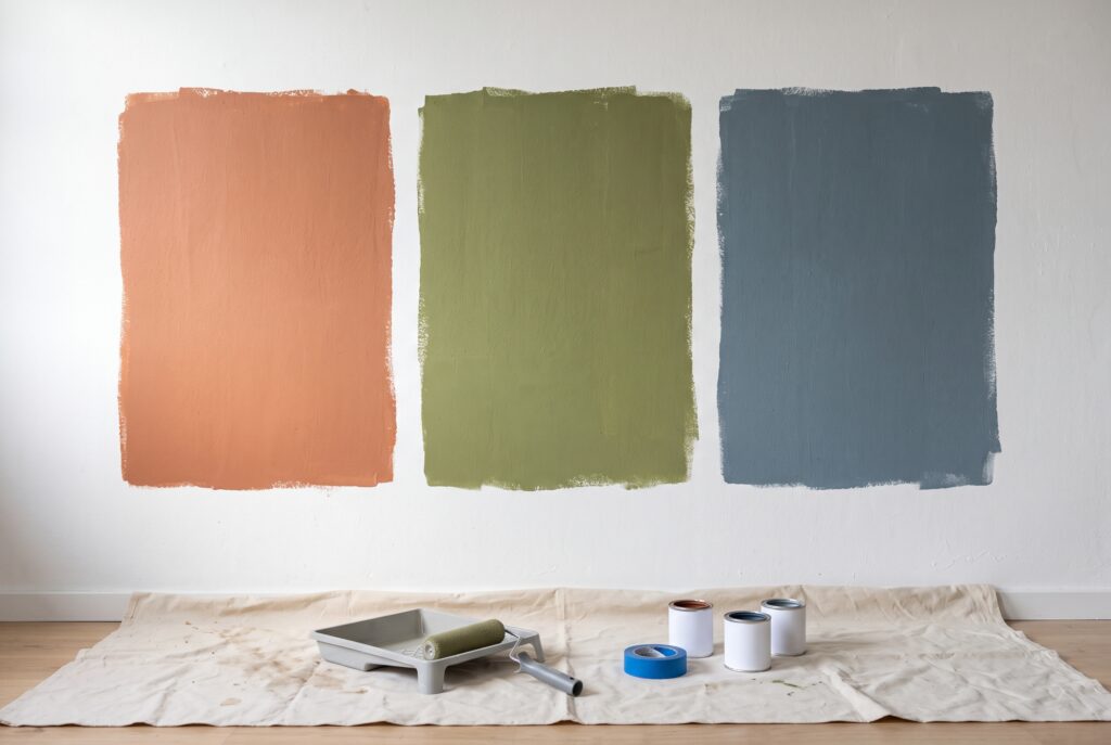 Pared con tres muestras grandes de pintura en tonos arcilla, verde oliva y azul grisáceo, con un rodillo pequeño y botes de muestra en el suelo