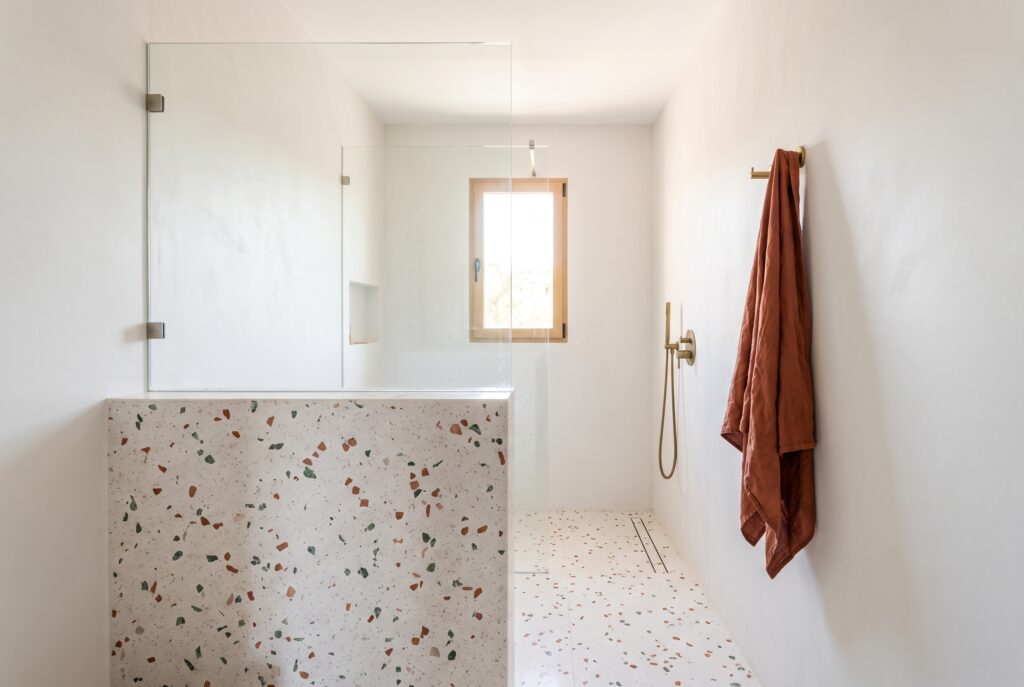 Ducha separada por un murete bajo de terrazo con un vidrio fijo encima, en un baño pequeño con luz natural y suelo de terrazo